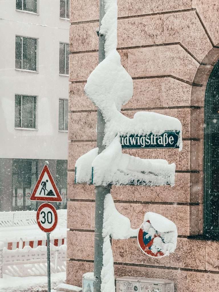 Snow-covered streets and buildings in Munich, with a picturesque winter landscape featuring frosted trees and a serene, white blanket of snow.