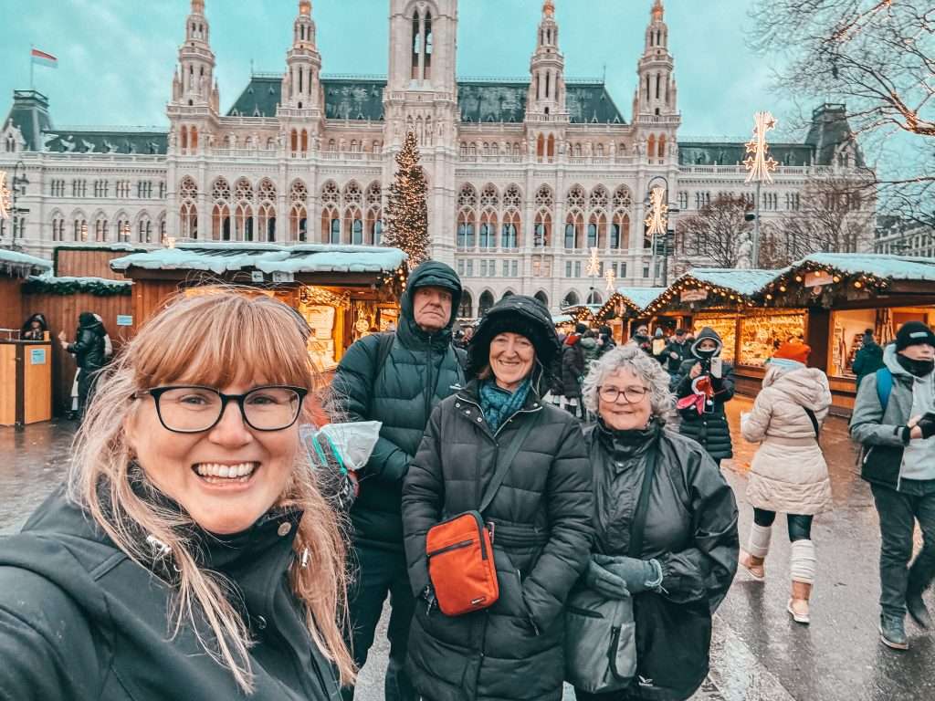 Our group at the Vienna Christmas Markets, warmly dressed in jackets and beanies, surrounded by festive holiday stalls and twinkling lights with snow softly falling