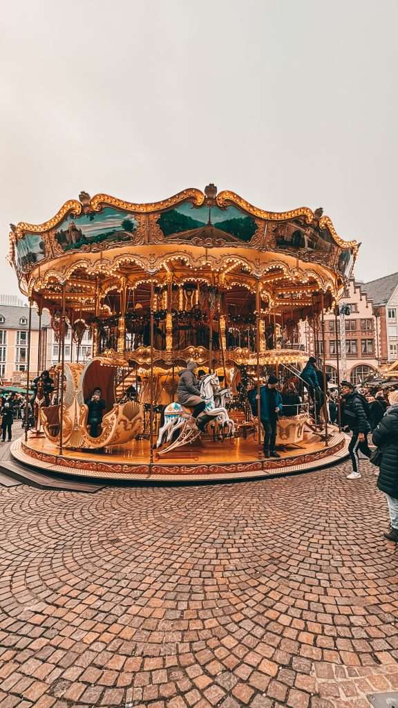 Frankfurt Christmas Markets bustling with holiday cheer, featuring traditional wooden stalls, festive lights, and seasonal decorations, with the historic architecture of the city in the background. Merry Go Round Seeds of Travel